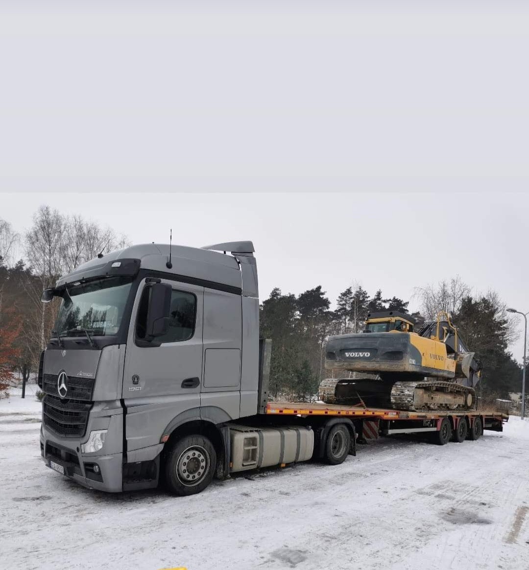 Szary ciągnik siodłowy Mercedes-Benz Actros przewozi na niskopodwoziowej platformie żółto-czarną koparkę Volvo w zimowej scenerii.