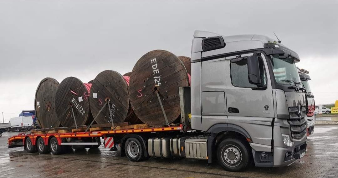 Srebrna ciężarówka Mercedes z naczepą przewozi duże, drewniane szpule z kablami w Chojnicach. Widoczne oznaczenia EDE/27 i EDE/28 na szpulach. Pochmurne niebo w tle.