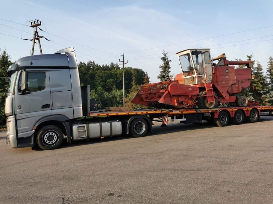 Szary ciągnik siodłowy z naczepą niskopodwoziową przewożący czerwony kombajn zbożowy. Widoczne drzewa i słupy wysokiego napięcia w tle.