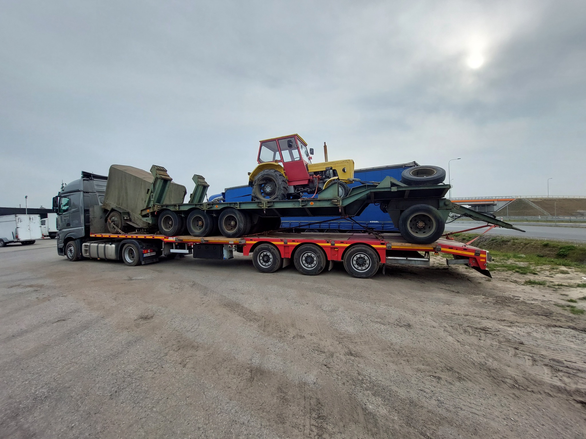 Ciężarówka transportuje traktor i sprzęt rolniczy na niskopodwoziowej naczepie, widok z boku na placu manewrowym.