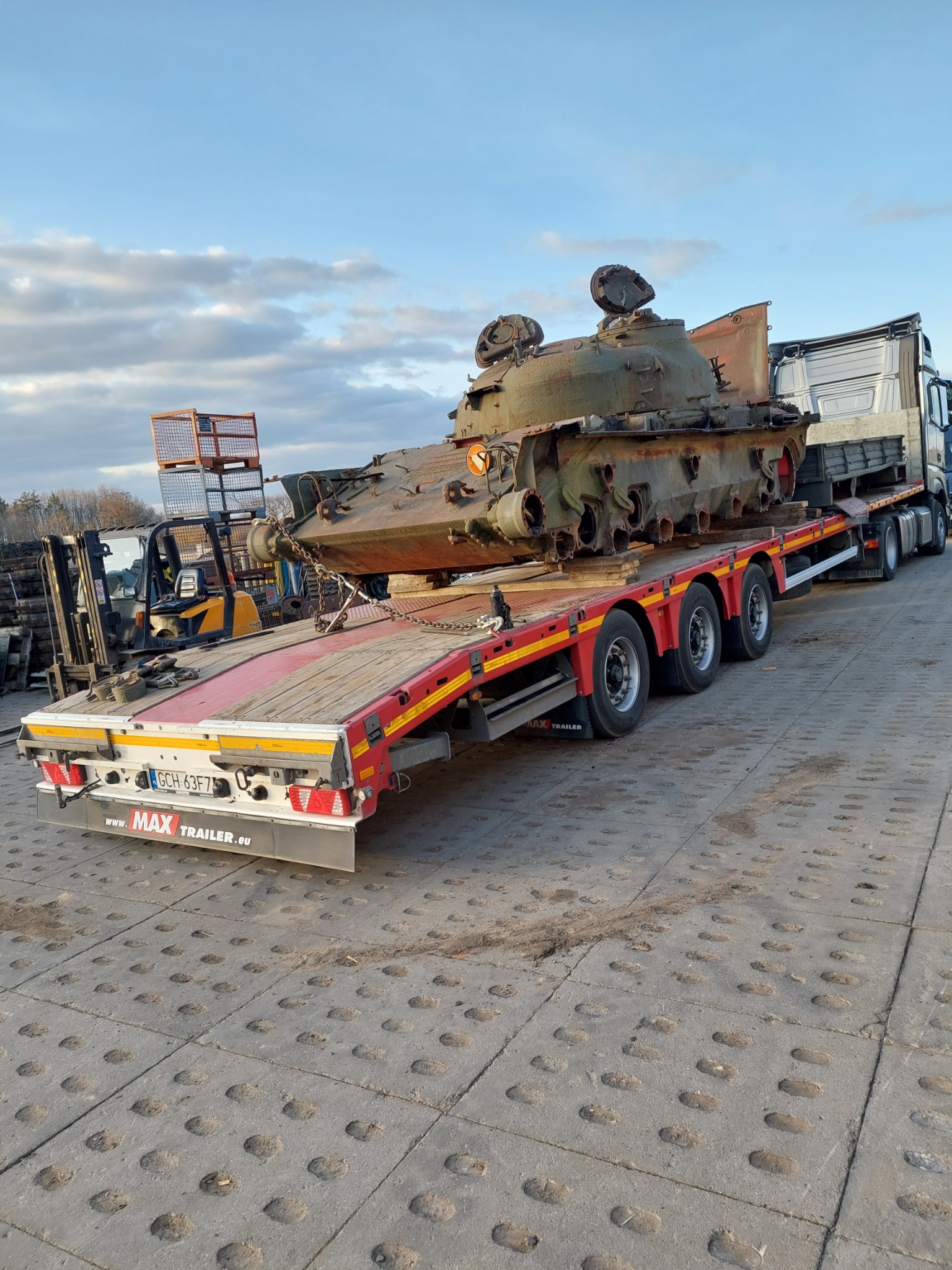 Transport czołgu T-55 na niskopodwoziowej platformie ciężarowej z żółtym wózkiem widłowym w tle, na placu z betonowej kostki.