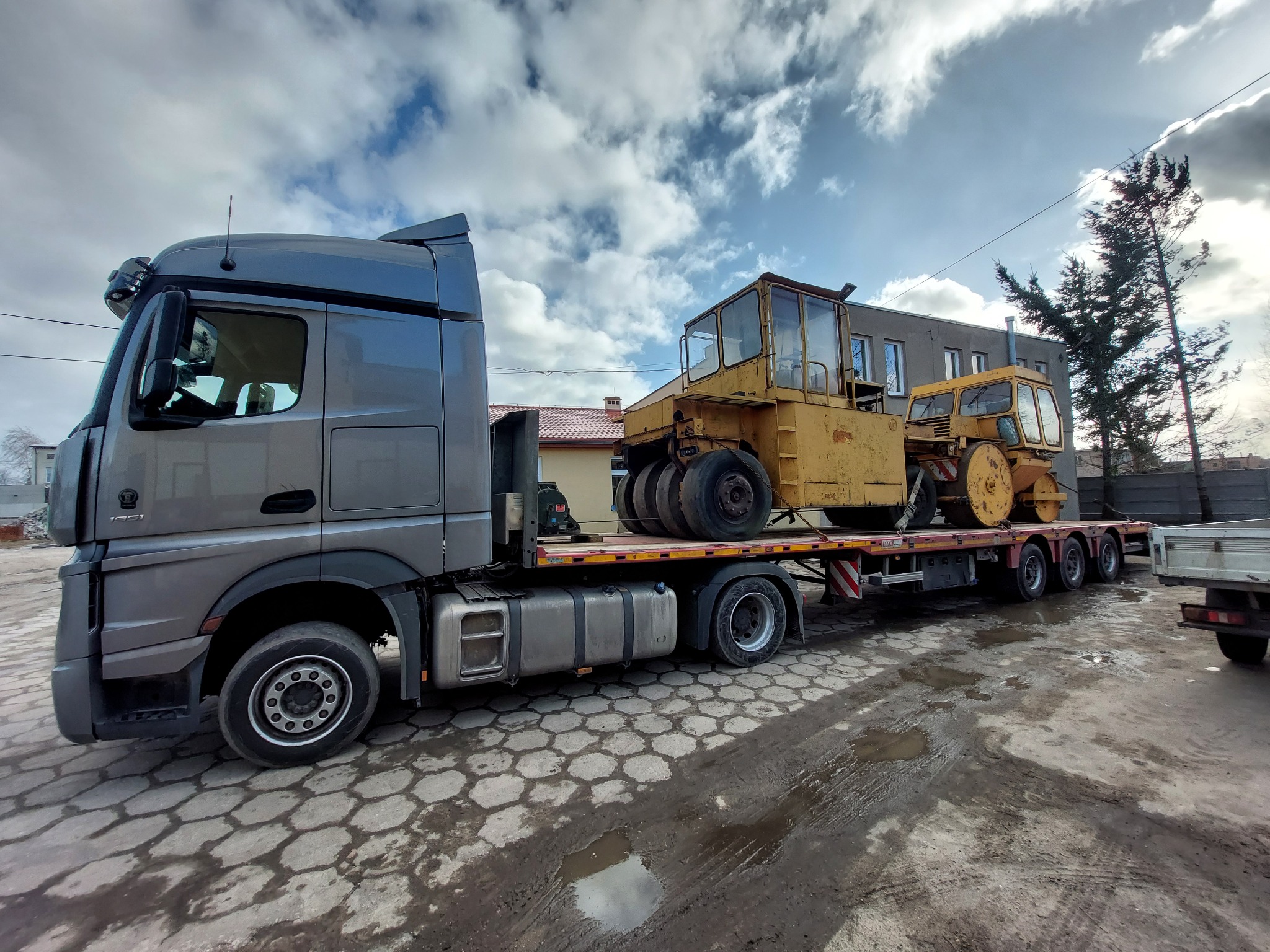 Szara ciężarówka z naczepą transportuje dwa żółte walce drogowe na tle pochmurnego nieba i budynków.