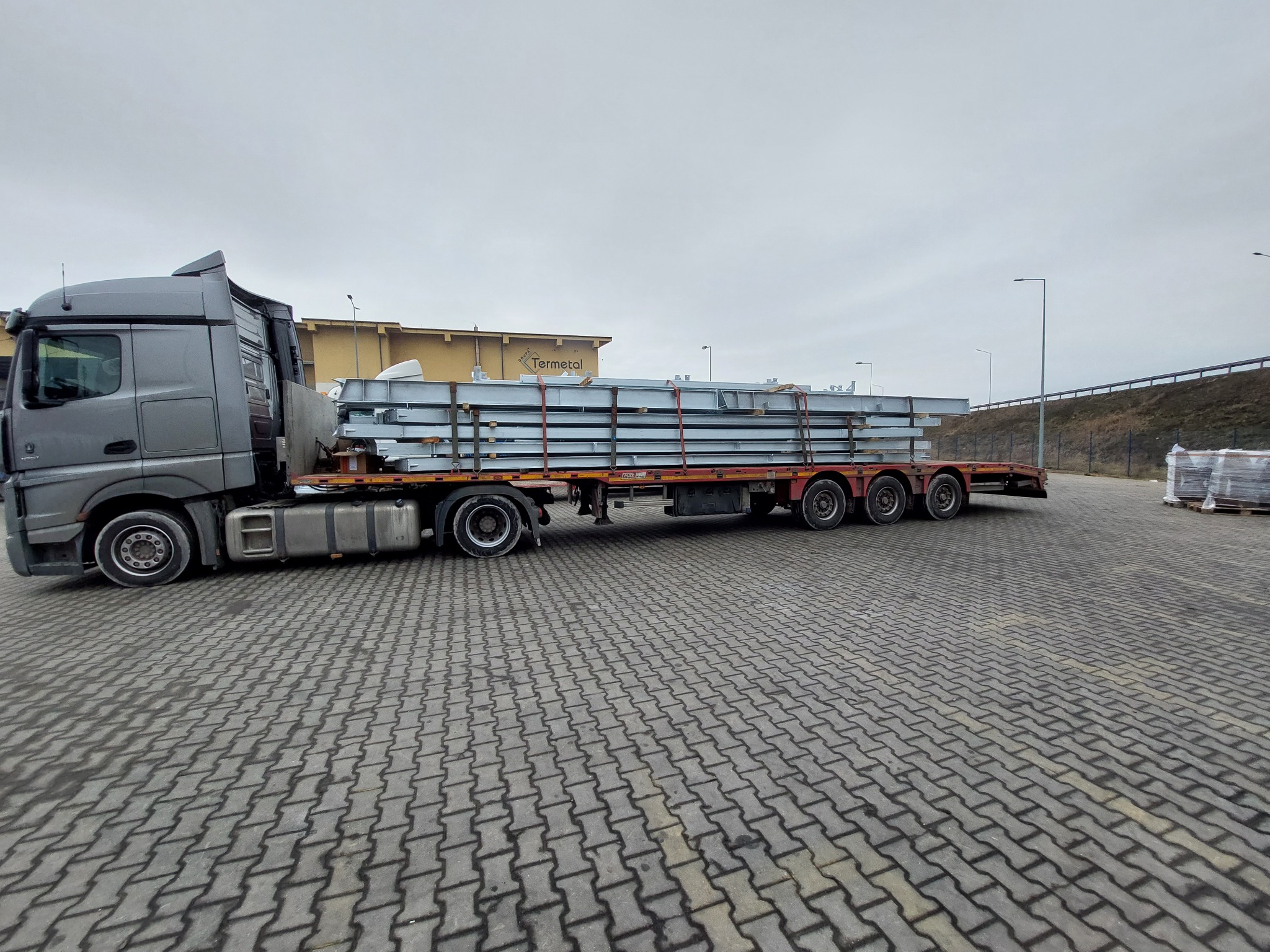 Szara ciężarówka z naczepą przewożąca stalowe konstrukcje, zabezpieczone czerwonymi pasami, na tle magazynu Termetal i pochmurnego nieba.