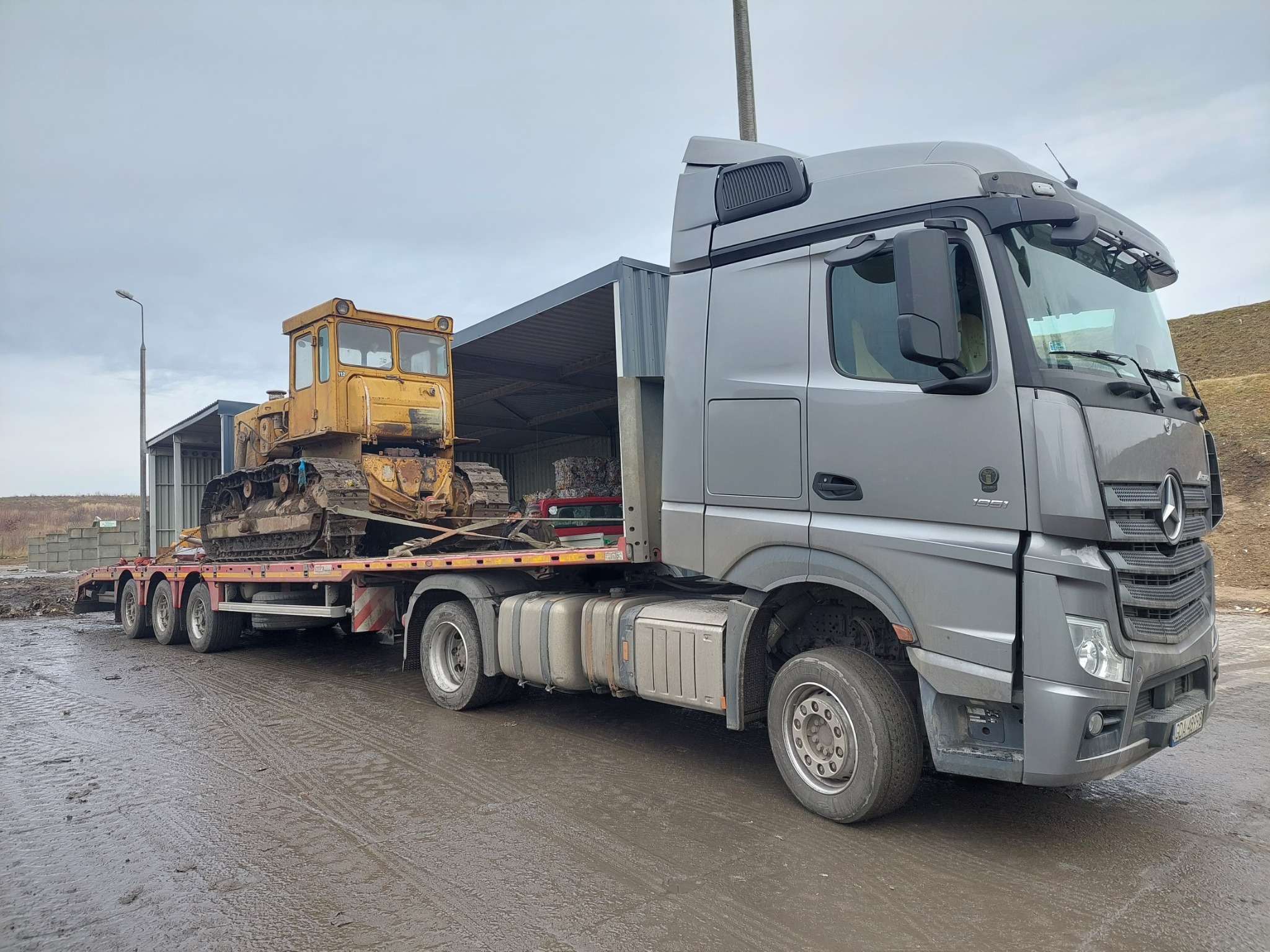 Szary ciągnik siodłowy z naczepą przewożący żółty, zabrudzony buldożer gąsienicowy na tle szarego nieba i hangaru.