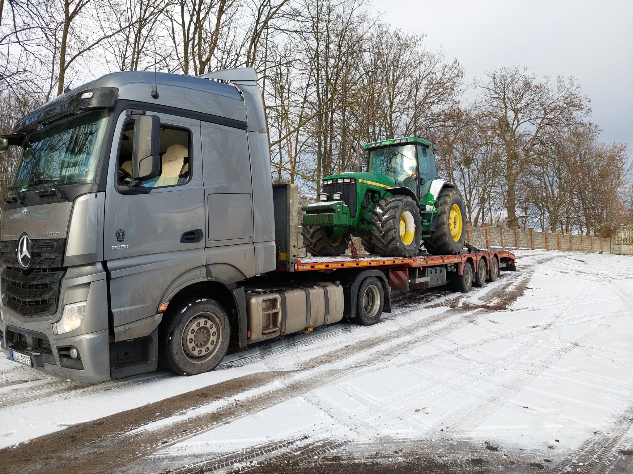 Szary ciągnik siodłowy Mercedes-Benz z naczepą przewożący zielony traktor John Deere na ośnieżonym placu, widoczne ślady opon