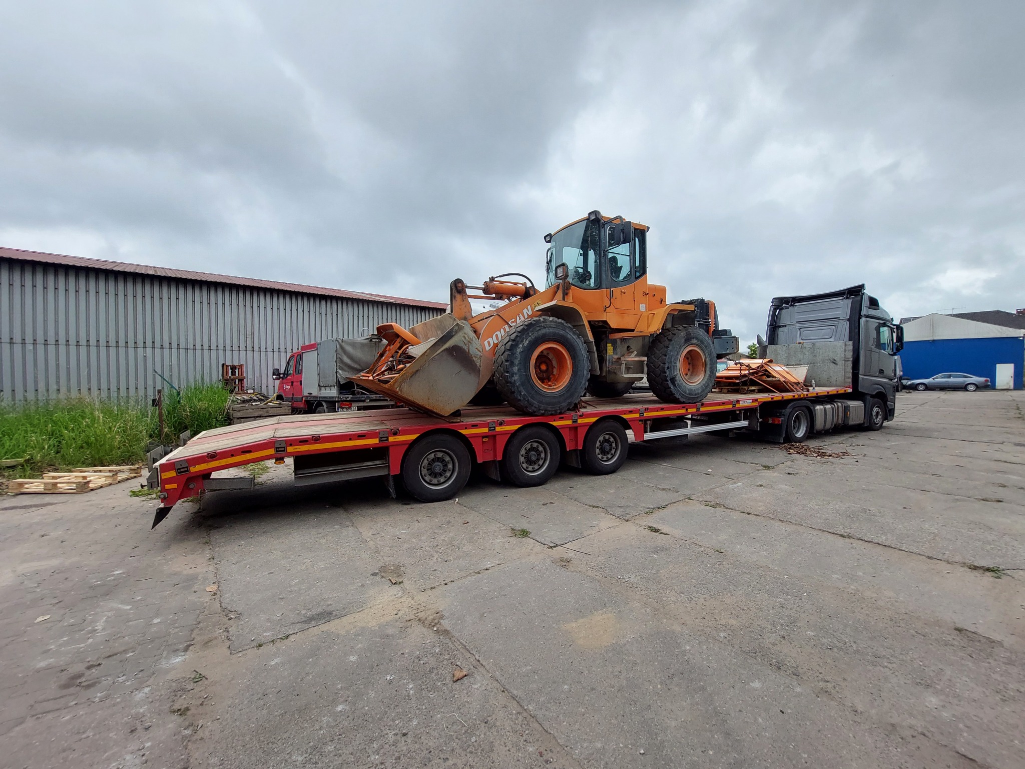 Pomarańczowa ładowarka kołowa Doosan przewożona na niskopodwoziowej platformie transportowej, widok z boku, w tle budynek magazynowy i pochmurne niebo.