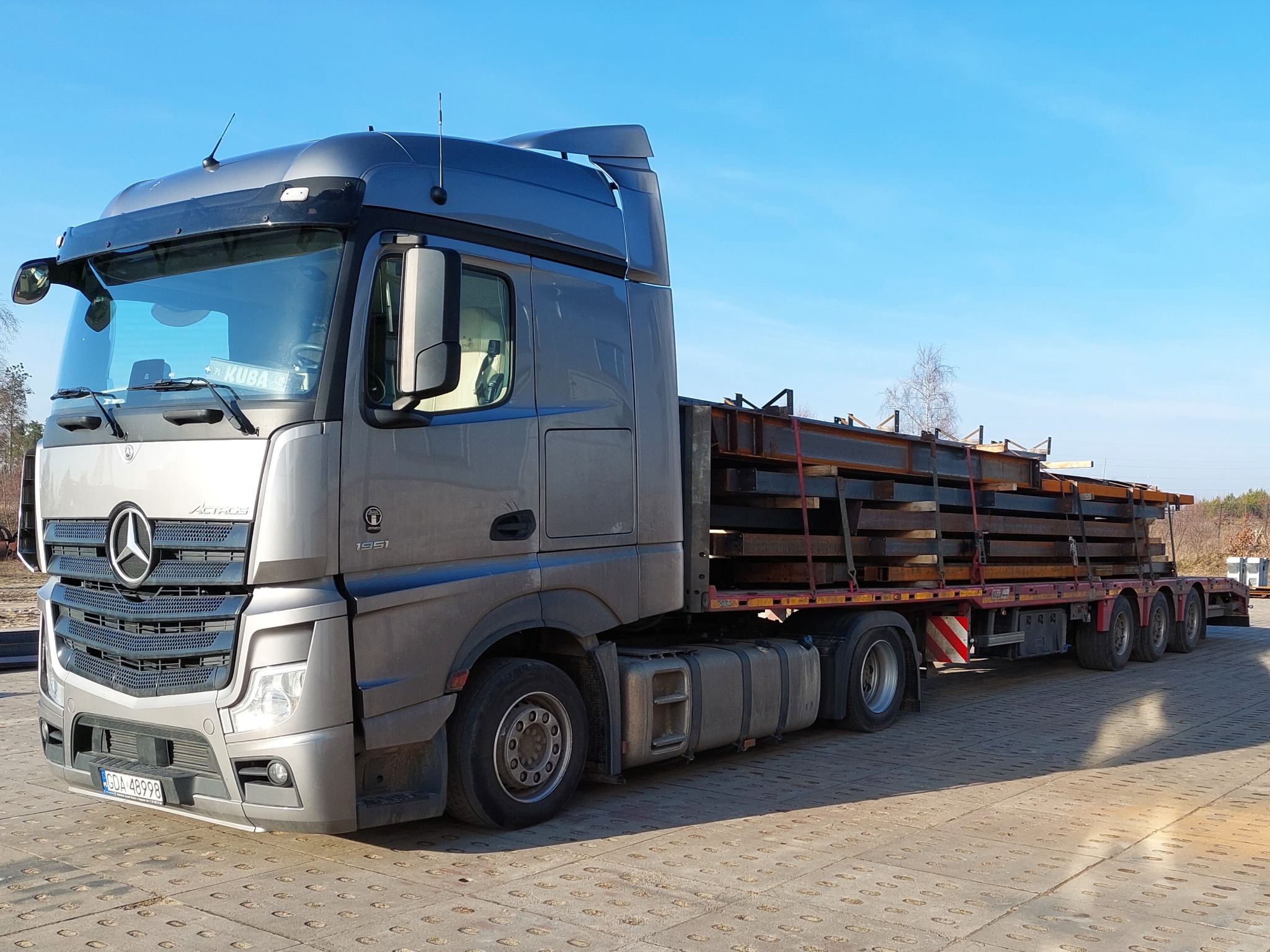 Szary ciągnik siodłowy Mercedes-Benz Actros z naczepą przewożącą stalowe elementy konstrukcyjne na tle błękitnego nieba.