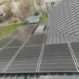 Panele słoneczne na grafitowym dachu domu jednorodzinnego, widok z góry na część paneli, okno dachowe i fragment ogrodu z elementami ogrodzenia.