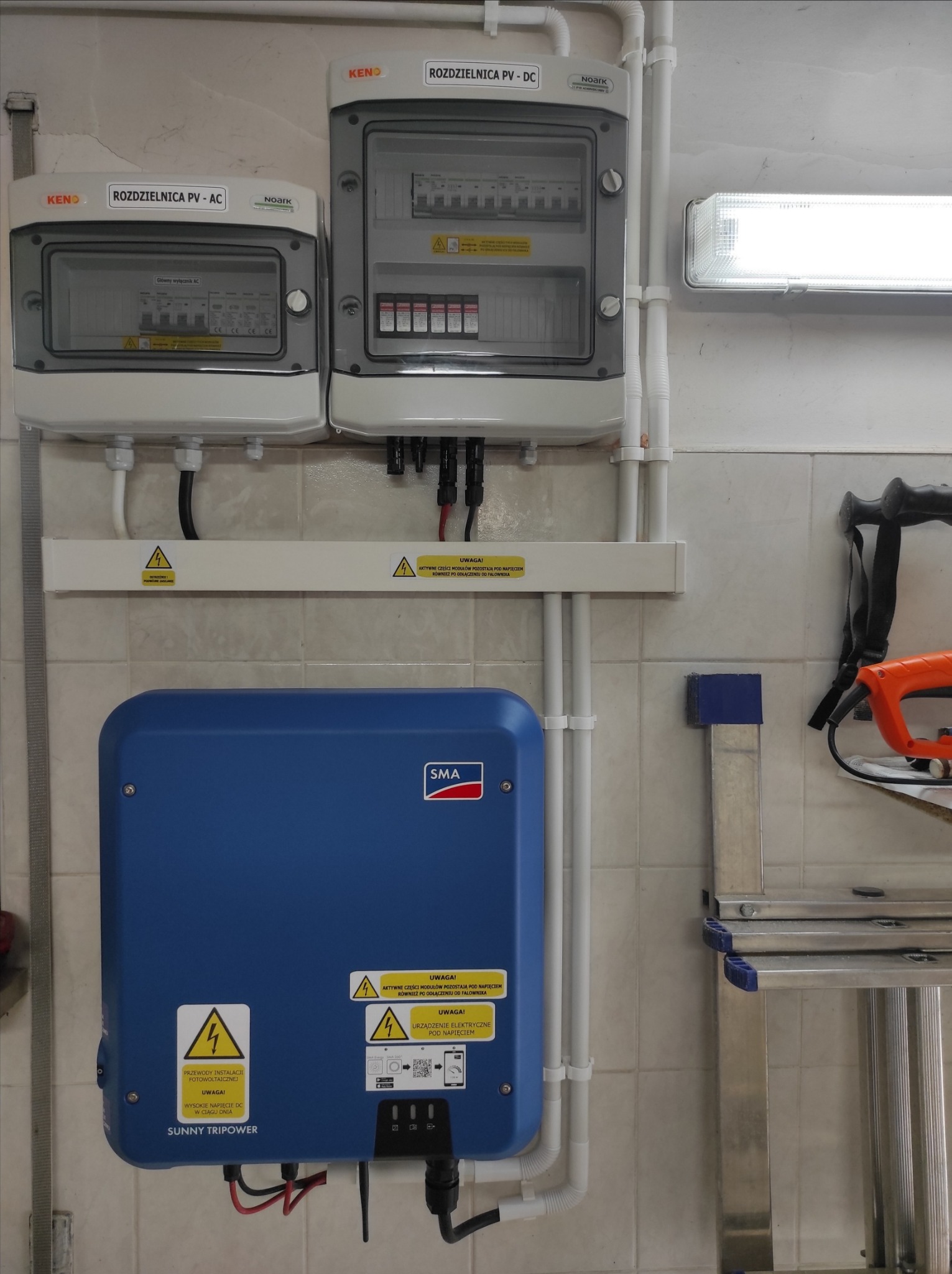 Rozdzielnice PV-AC i PV-DC marki KENO i Noark, niebieski falownik SMA Sunny Tripower oraz fragment drabiny aluminiowej i narzędzia na tle ściany z płytek