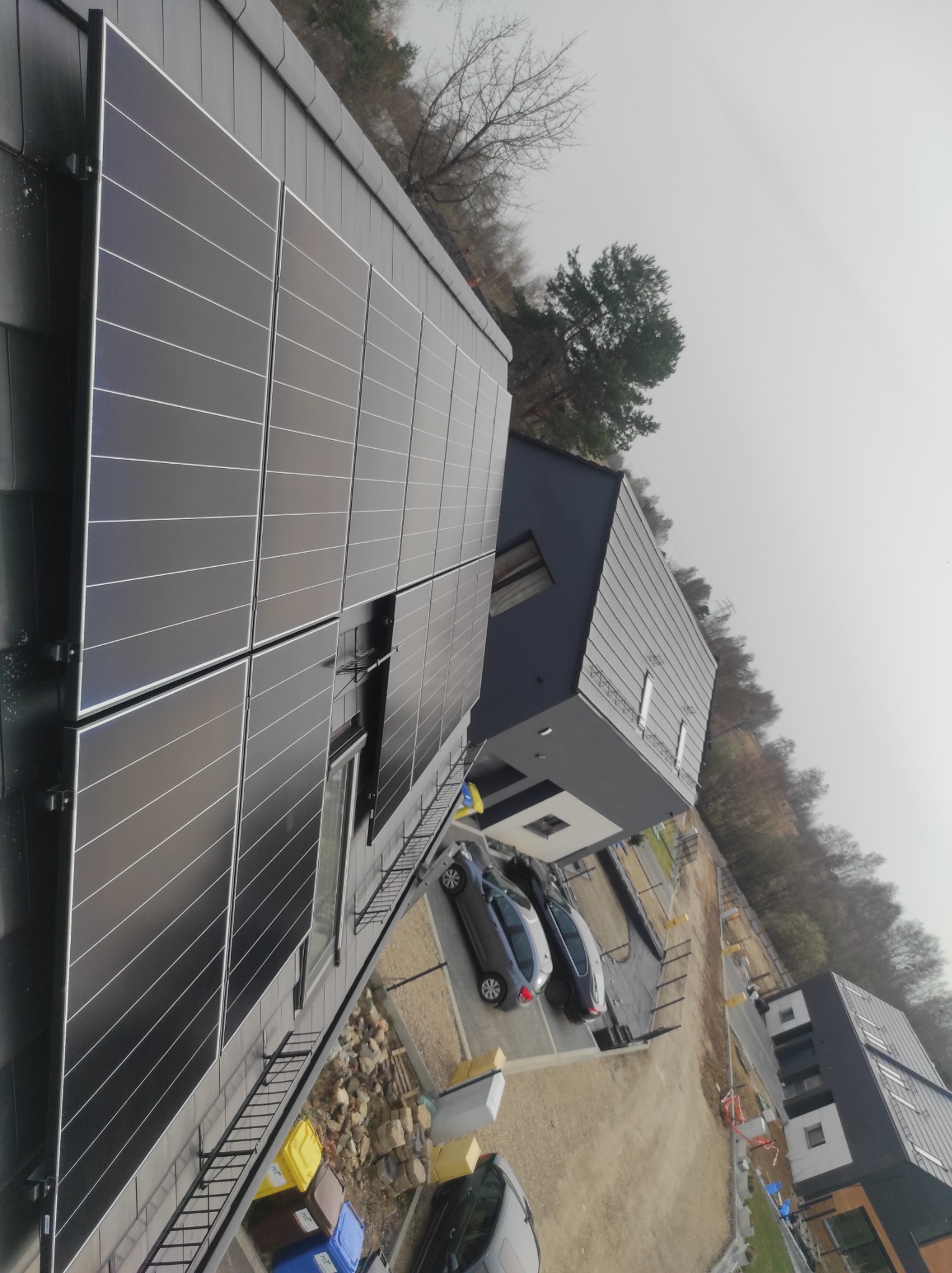 Nowoczesny dom z panelami słonecznymi na dachu i elewacji, widok z góry, samochody na podjeździe, częściowo zagospodarowany teren.