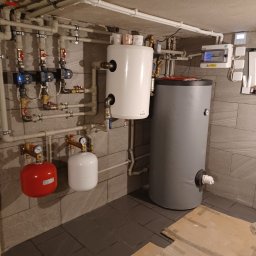 Piotrowski - Złożona instalacja hydrauliczna z dwoma zbiornikami, pompami obiegowymi i rozdzielnicą elektryczną na tle szarych płytek ściennych i podłogowych.