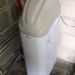 Piotrowski - Nowoczesny zmiękczacz wody USTM w piwnicy, widoczne rury instalacyjne, narzędzia hydrauliczne i kabel zasilający na podłodze z płytek.