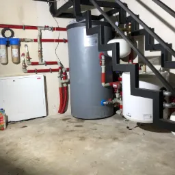 Kotłownia z szarym zbiornikiem akumulacyjnym, białym zbiornikiem wyrównawczym, filtrami wody i instalacją hydrauliczną z rur miedzianych, widoczna spod metalowych schodów.