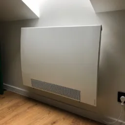 Biały, płaski grzejnik elektryczny marki Atlantic zamontowany na ścianie w pokoju na poddaszu ze skośnym sufitem i świetlikiem, widoczny kabel zasilający i gniazdko elektryczne.