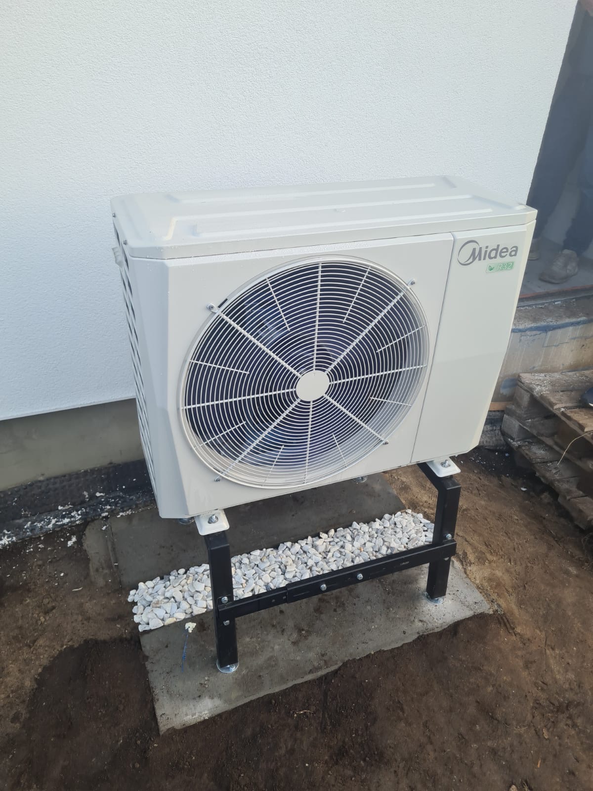 Biała zewnętrzna jednostka klimatyzacji marki Midea na czarnym, metalowym stelażu, umieszczona na betonowej podstawie z białym żwirem, przy białej ścianie budynku. Widoczne elementy instalacji...