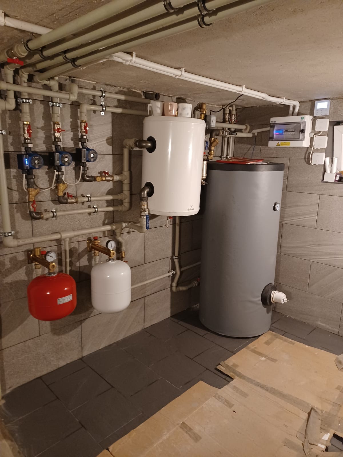 Złożona instalacja hydrauliczna z dwoma zbiornikami, pompami obiegowymi i rozdzielnicą elektryczną na tle szarych płytek ściennych i podłogowych.
