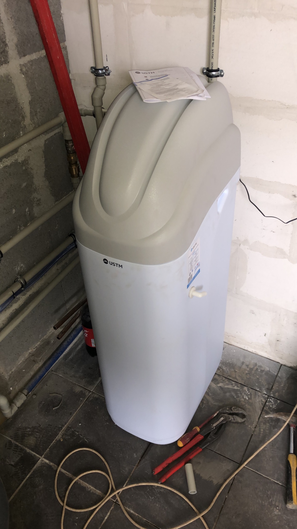 Nowoczesny zmiękczacz wody USTM w piwnicy, widoczne rury instalacyjne, narzędzia hydrauliczne i kabel zasilający na podłodze z płytek.
