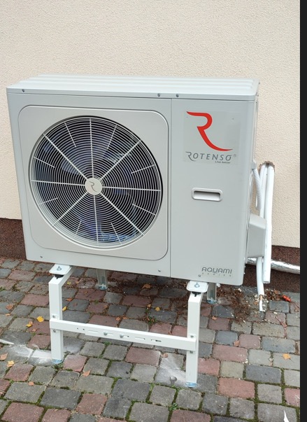 Zewnętrzna jednostka klimatyzacji Rotenso Aquami, zamontowana na białym stelażu, z widocznymi przewodami przy elewacji budynku.