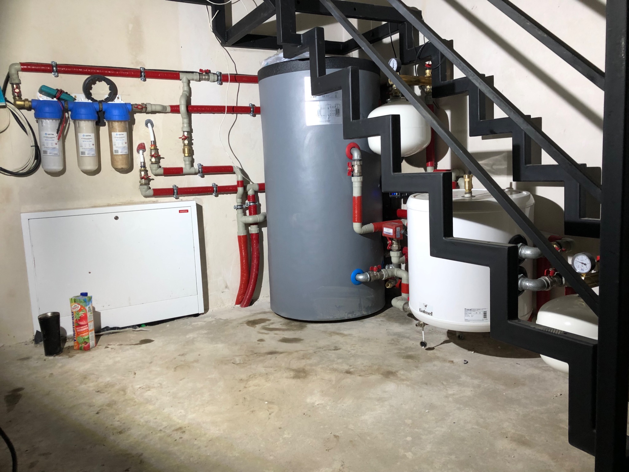 Kotłownia z szarym zbiornikiem akumulacyjnym, białym zbiornikiem wyrównawczym, filtrami wody i instalacją hydrauliczną z rur miedzianych, widoczna spod metalowych schodów.
