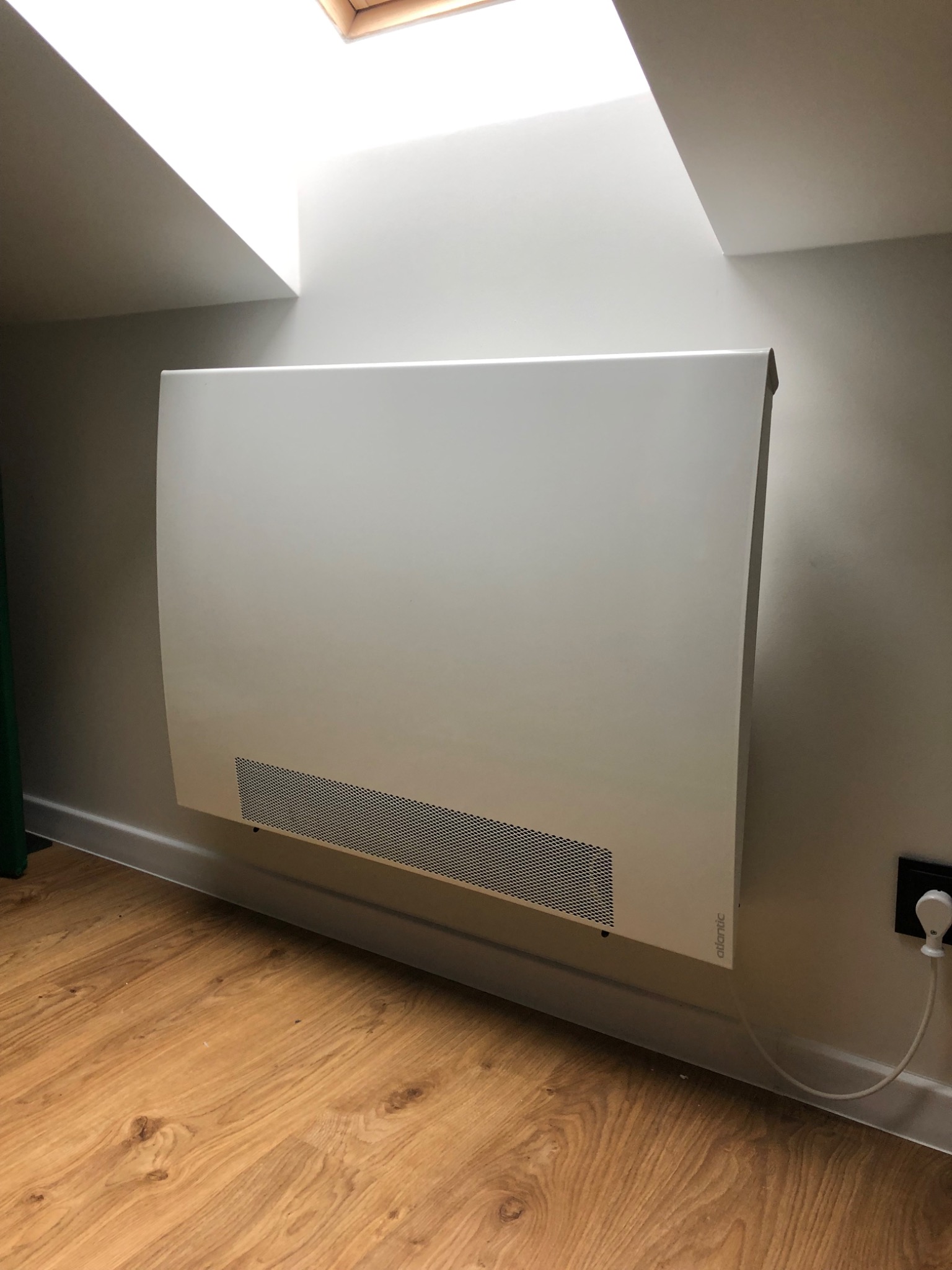 Biały, płaski grzejnik elektryczny marki Atlantic zamontowany na ścianie w pokoju na poddaszu ze skośnym sufitem i świetlikiem, widoczny kabel zasilający i gniazdko elektryczne.