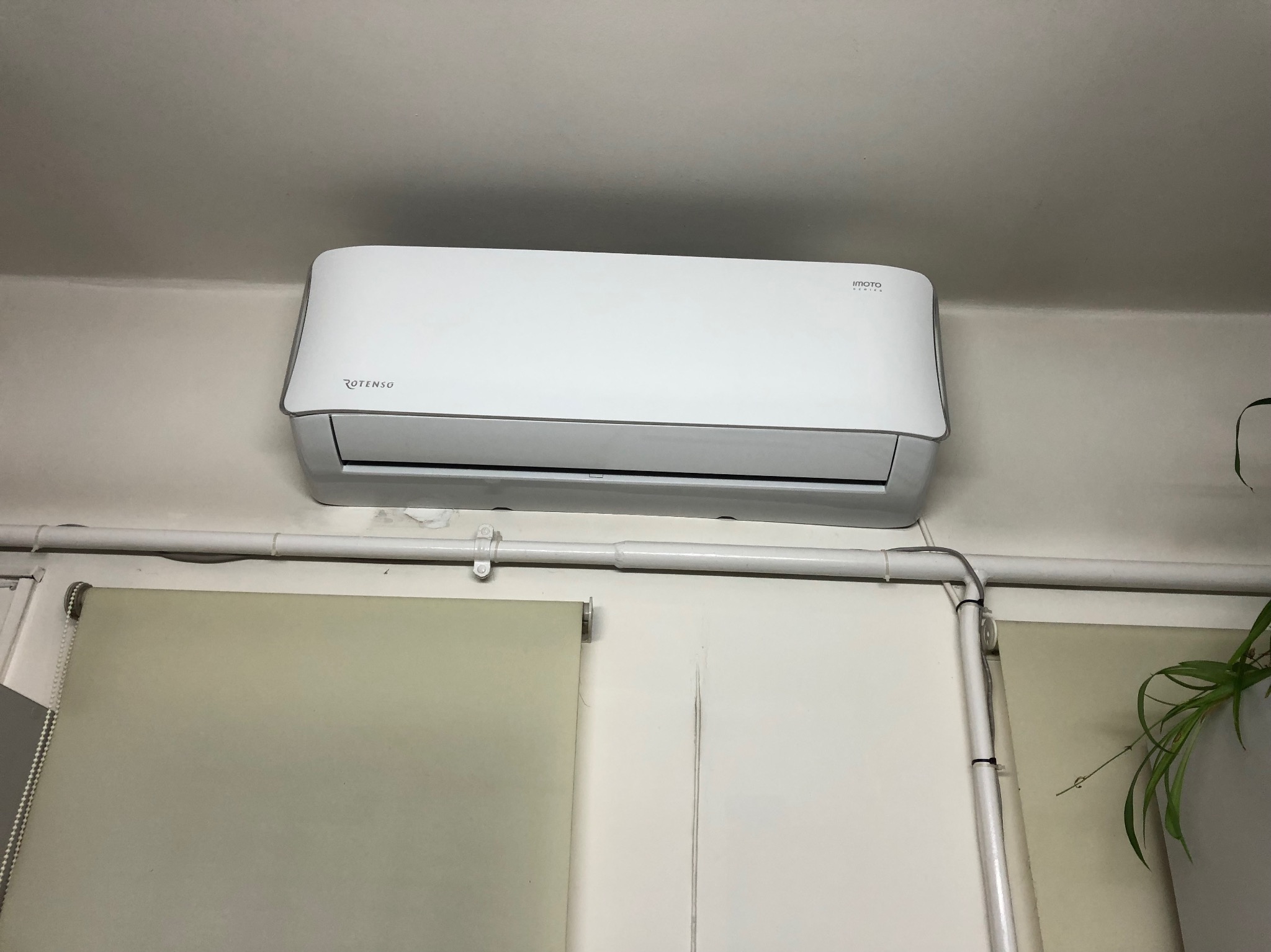 Biała jednostka klimatyzacji Rotenso zamontowana na ścianie nad roletą okienną, widoczne rury instalacyjne przy ścianie i fragment rośliny doniczkowej.