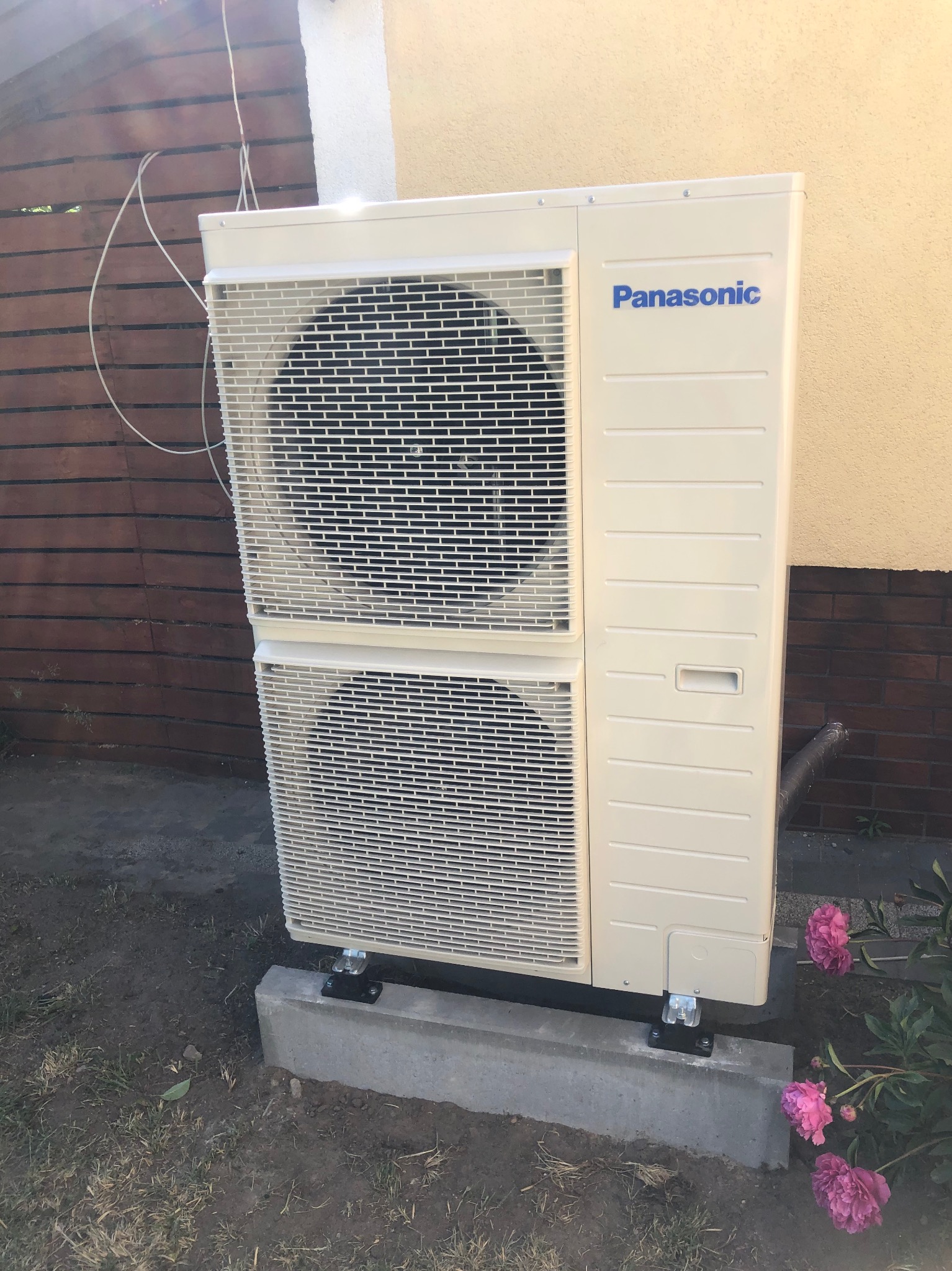 Zewnętrzna jednostka klimatyzacji Panasonic zamontowana na betonowej podstawie, widoczne przewody i kwiaty piwonii w tle.