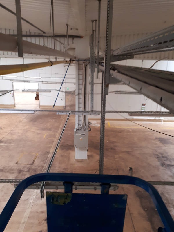 Widok z góry na dużą halę z widocznymi instalacjami pod sufitem: koryta kablowe, rury i skrzynka elektryczna na słupie. Perspektywa z podnośnika koszowego.