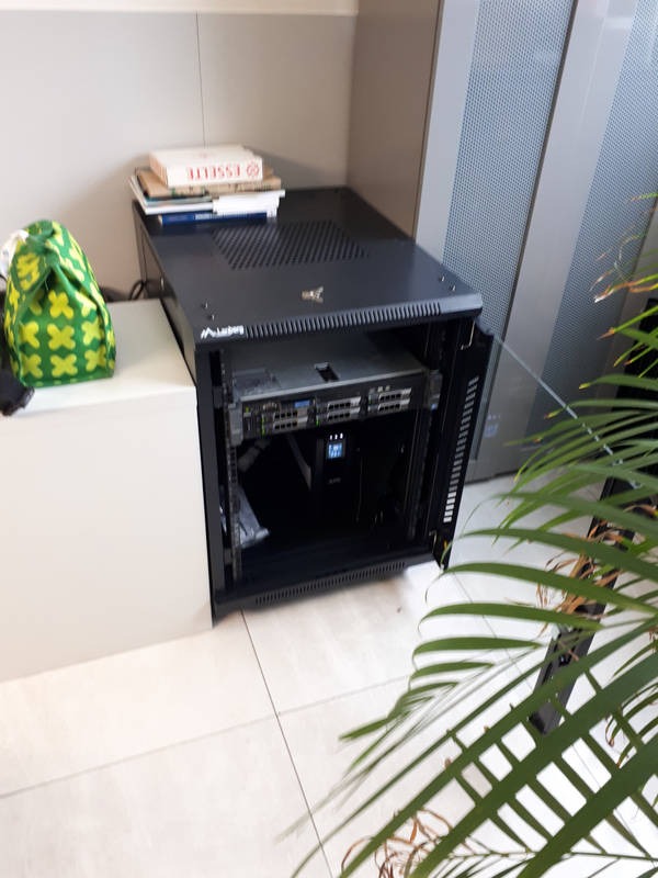 Otwarta szafa rack z serwerem, UPS i okablowaniem w pomieszczeniu biurowym, torba z zielonym wzorem obok, książki na górze szafy.