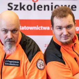 Portret dwóch mężczyzn w pomarańczowych polarach z logo 'Pogotowie Szkoleniowe' na tle baneru z napisem 'Szkoleniowe ratownictwa'