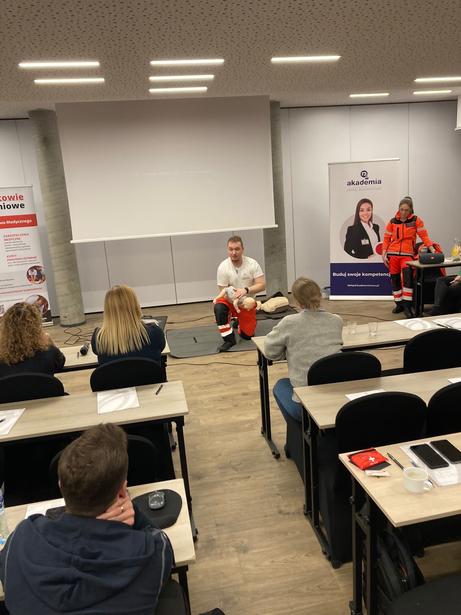 Instruktor demonstruje resuscytację krążeniowo-oddechową na manekinie niemowlęcia podczas zajęć z pierwszej pomocy dla grupy osób w sali konferencyjnej, obok widoczny baner reklamowy z logiem firmy...