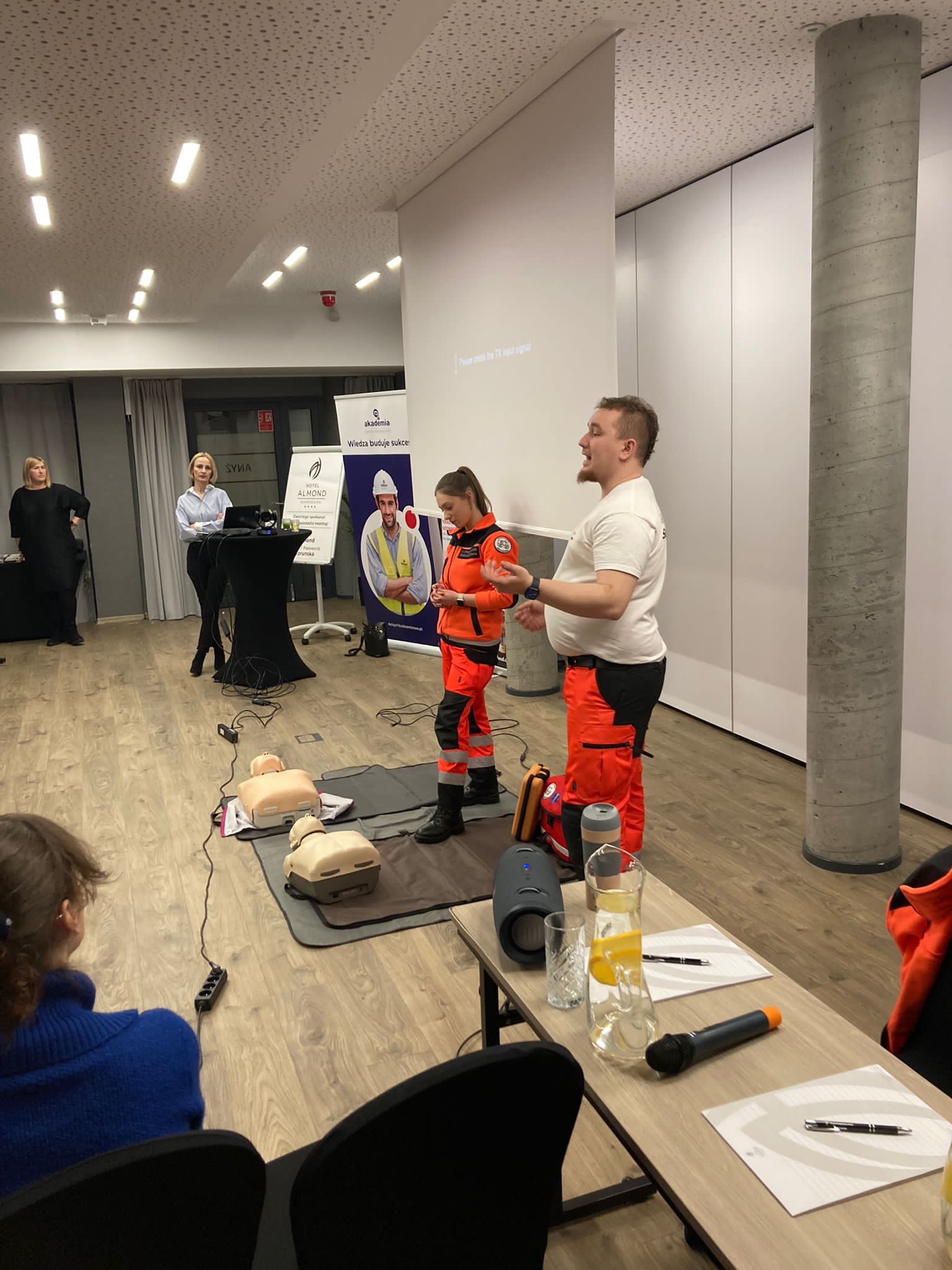 Instruktor w pomarańczowym kombinezonie demonstruje resuscytację na fantomie podczas szkolenia, obok stoi drugi instruktor, w tle tablica z logo firmy szkoleniowej.