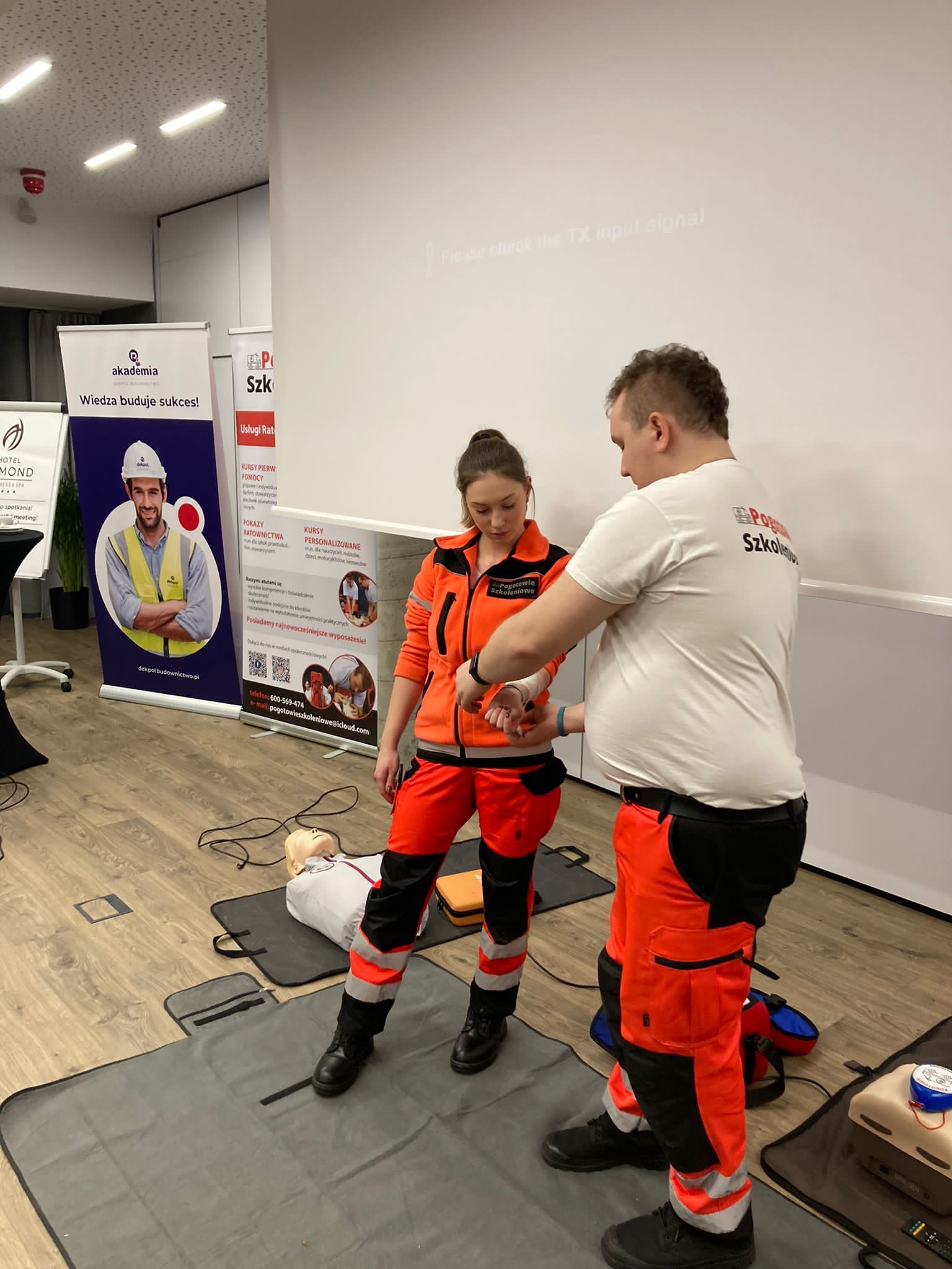 Instruktor w białej koszulce demonstruje technikę zakładania opaski uciskowej na przedramieniu kursantki ubranej w strój ratowniczy, w tle manekin do ćwiczeń i roll-upy reklamowe.