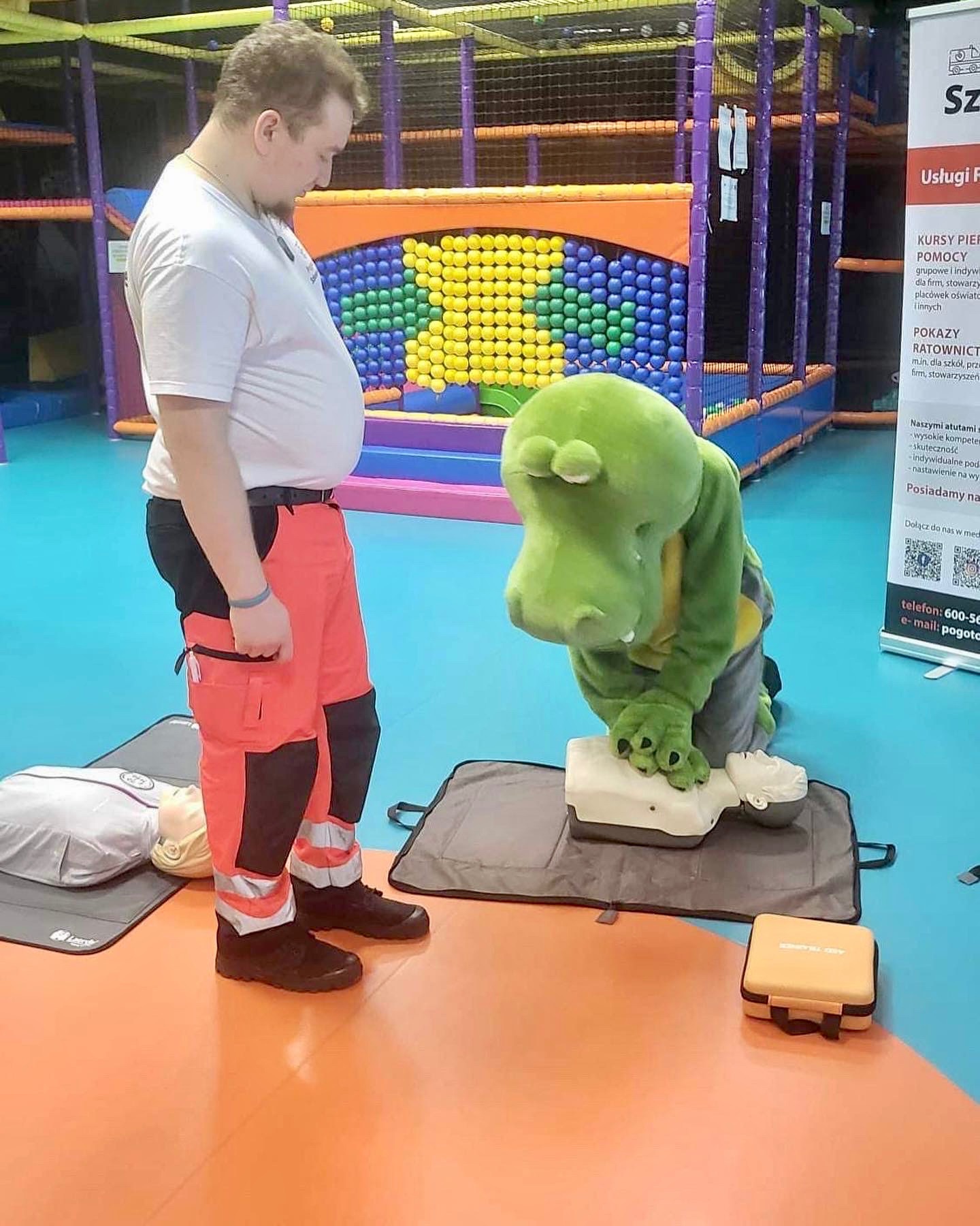 Instruktor w stroju krokodyla demonstruje resuscytację krążeniowo-oddechową na fantomie, obok stoi ratownik medyczny w pomarańczowych spodniach na tle sali zabaw.