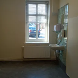 Wnętrze łazienki z umywalką, grzejnikiem pod oknem z żaluzjami i ciemną podłogą z płytek. Na zewnątrz widoczny samochód osobowy.