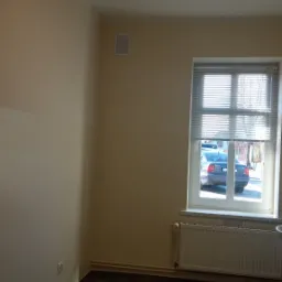 Pomalowane na beżowo wnętrze pokoju z ciemną, błyszczącą podłogą z płytek, biały grzejnik pod oknem z żaluzjami, widoczny fragment samochodu za oknem.