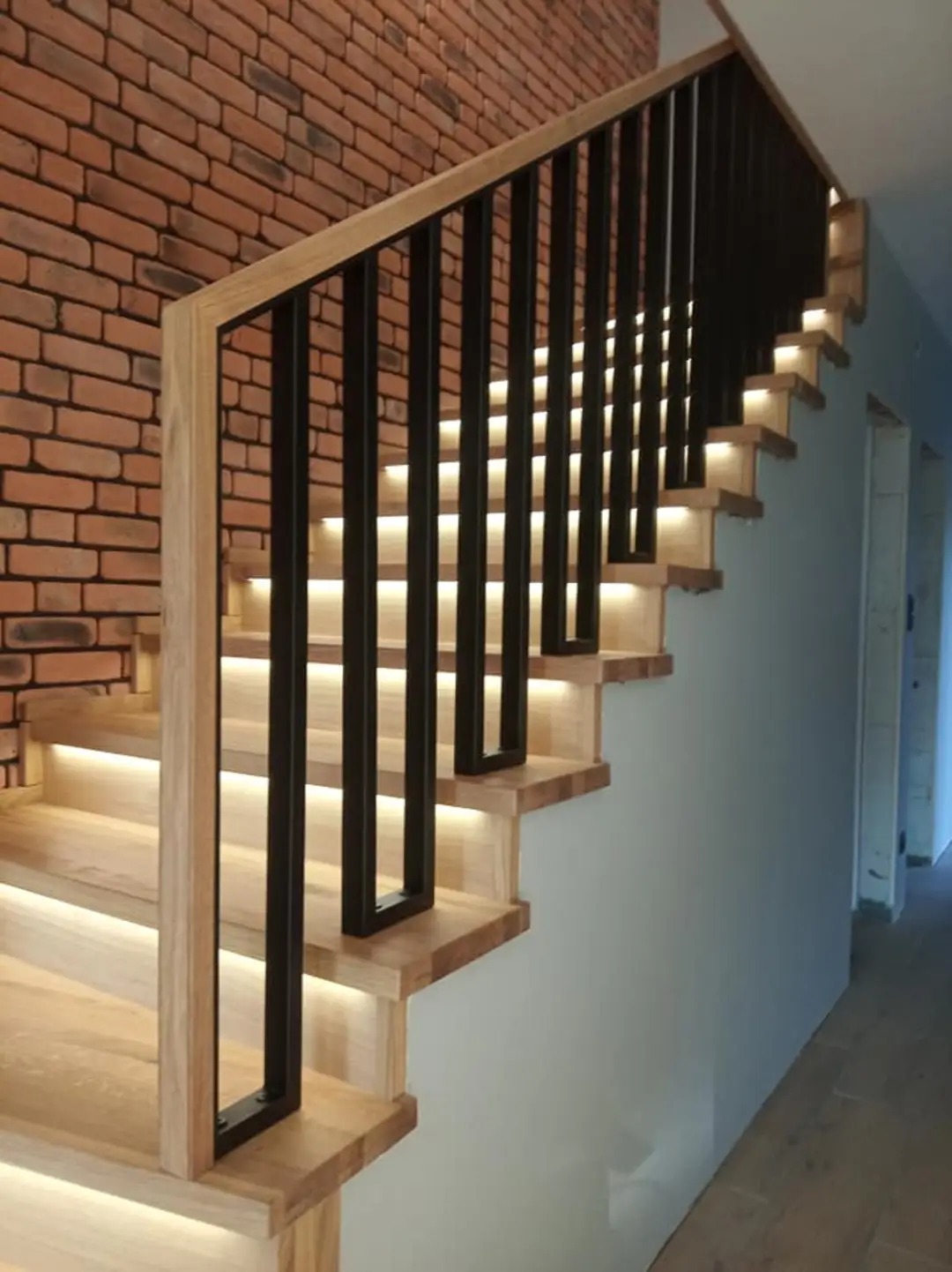 Nowoczesne schody z drewnianymi stopniami podświetlonymi ledami, czarne metalowe barierki, ściana z cegły w tle.