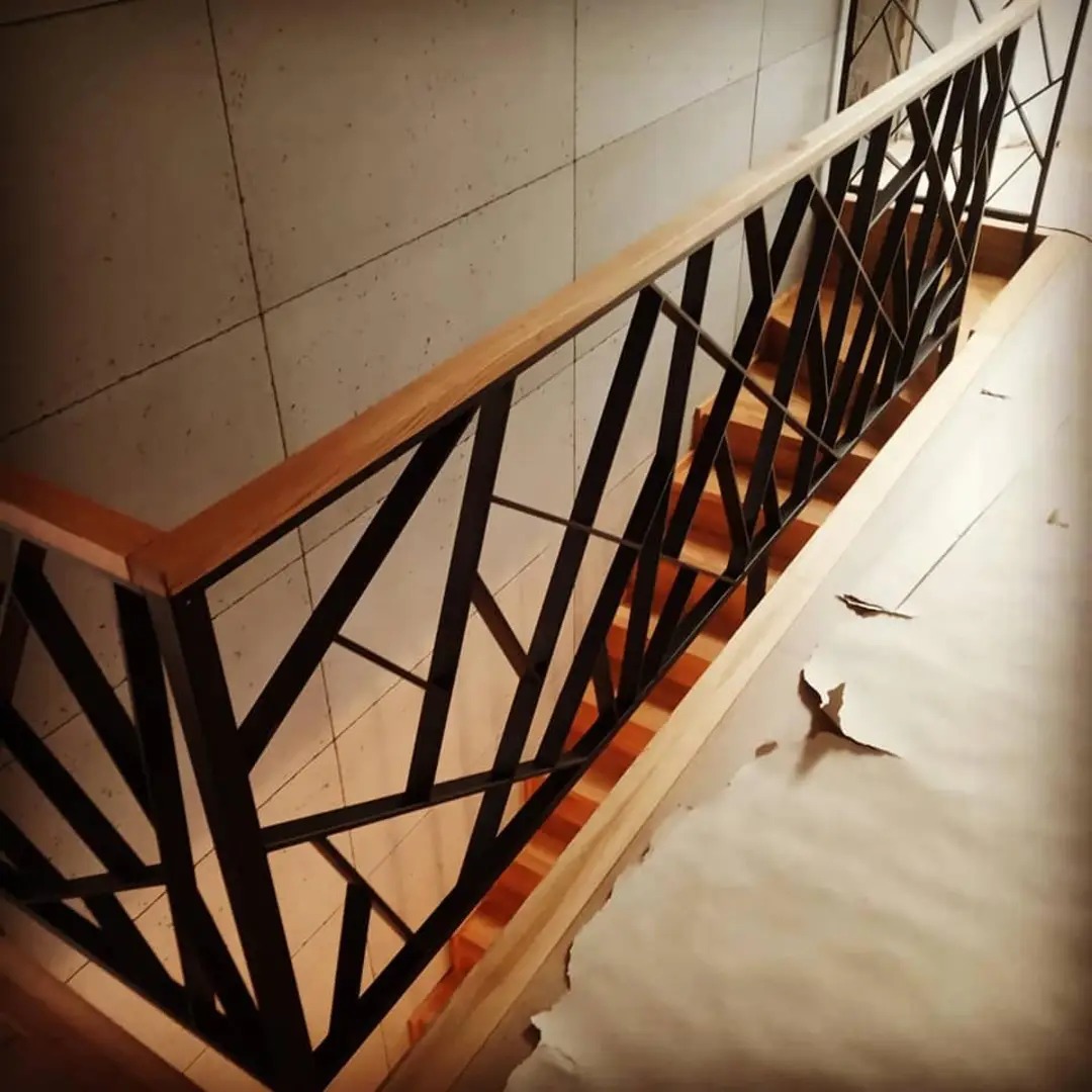 Nowoczesne schody z metalową balustradą o geometrycznym wzorze i drewnianym poręczą, widoczne z góry, z elementami wykończeniowymi na podłodze.