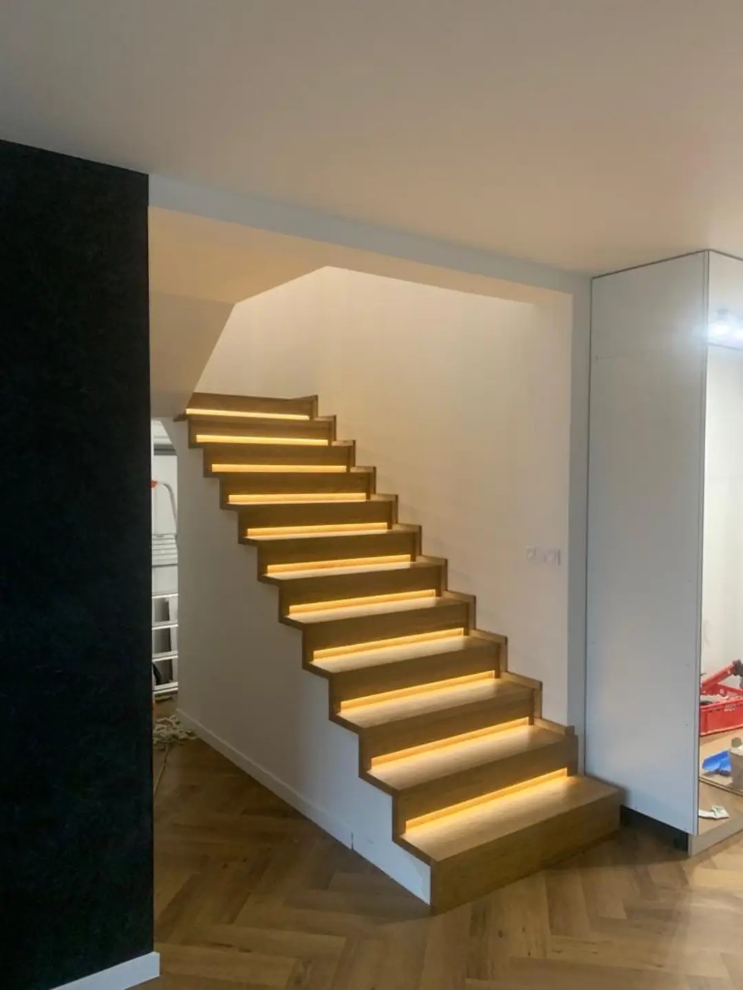 Nowoczesne, drewniane schody z podświetleniem LED na każdym stopniu, widok z dołu, parkiet w jodełkę na podłodze, czarna ściana z lewej strony.