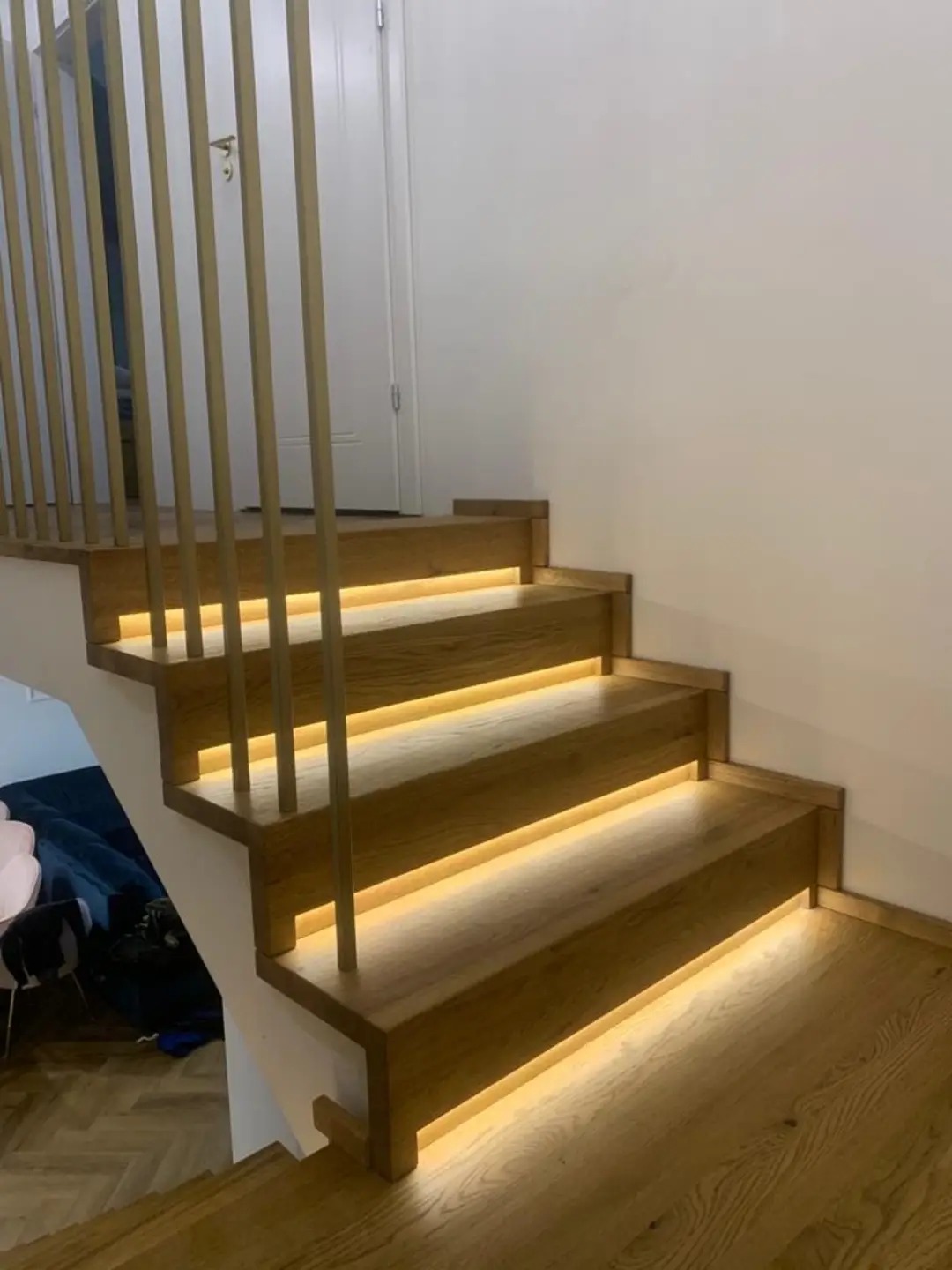 Nowoczesne schody drewniane z podświetleniem LED na każdym stopniu i minimalistyczną balustradą z pionowych, złotych prętów.