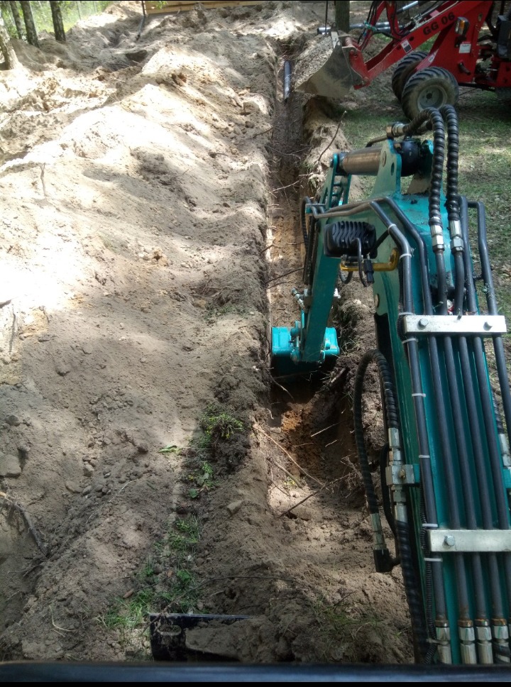 Wąski wykop w ziemi z widoczną rurą, wykonywany małą koparką w słoneczny dzień, z fragmentem większej maszyny w tle.