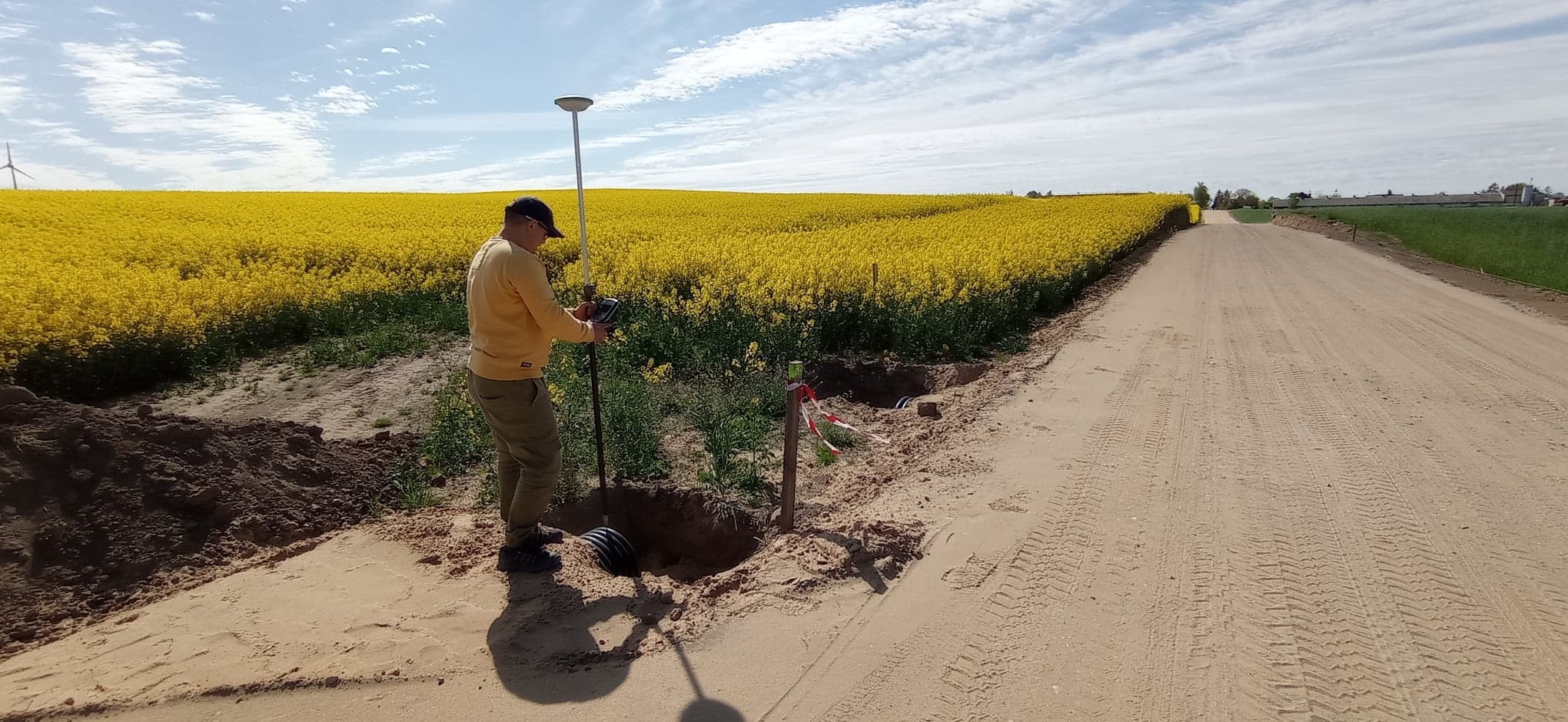 Pracownik w żółtej bluzie używa instrumentu pomiarowego GPS nad wykopem przy piaszczystej drodze, z polem kwitnącego rzepaku w tle.
