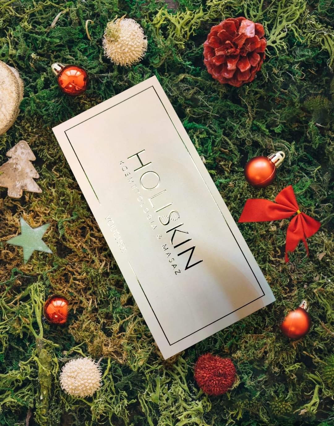 Karta podarunkowa Holiskin Kosmetologia i Masaż, otoczona świątecznymi dekoracjami na tle z mchu.