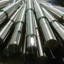 Stos polerowanych, metalowych wałków z wyfrezowanymi otworami i gwintowanymi końcówkami, ułożonych pod kątem, w tle widoczna hala produkcyjna.