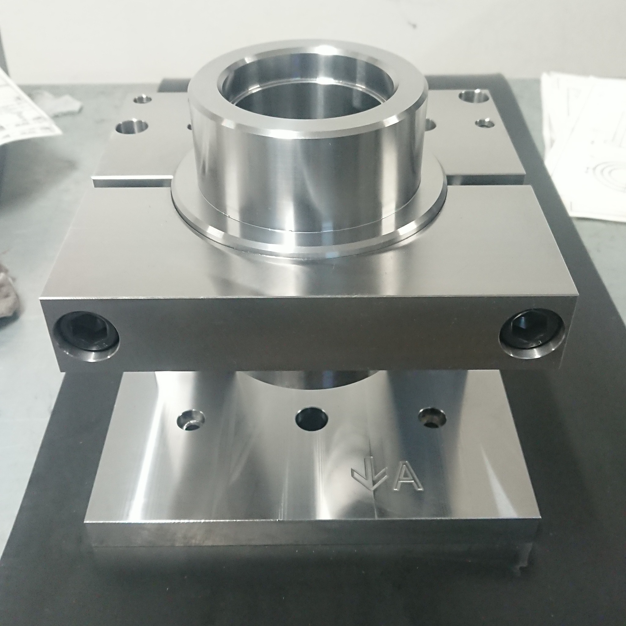 Precyzyjnie wykonany, metalowy element z widocznymi śladami obróbki skrawaniem, z centralnym cylindrycznym otworem i wygrawerowanym znakiem strzałki w dół oraz literą A.