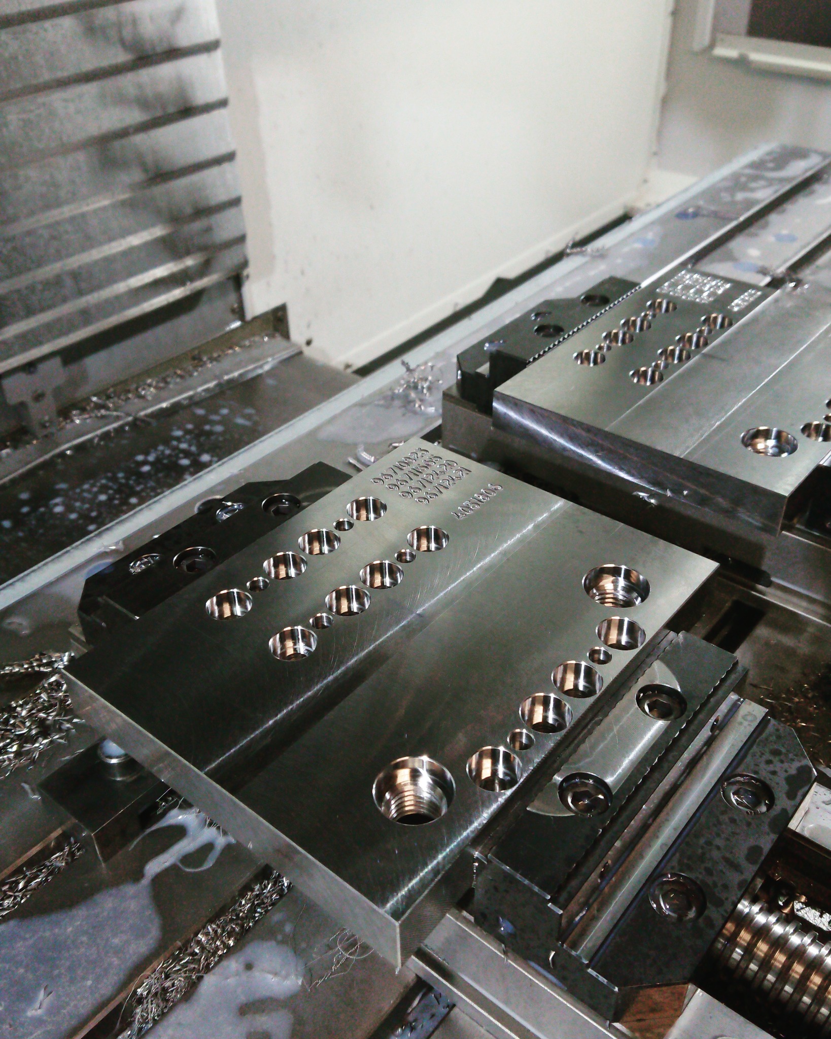 Dwie metalowe płyty zamocowane w uchwycie obrabiarki CNC, z wywierconymi otworami o różnych średnicach i wygrawerowanymi numerami seryjnymi, widoczne wióry i płyn chłodzący.