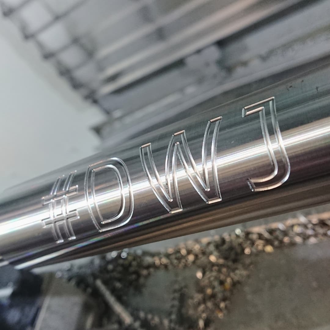 Metalowy wałek z precyzyjnie wygrawerowanym napisem '#DWJ', widoczne ślady obróbki i opiłki metalu, wykonane w technologii CNC.
