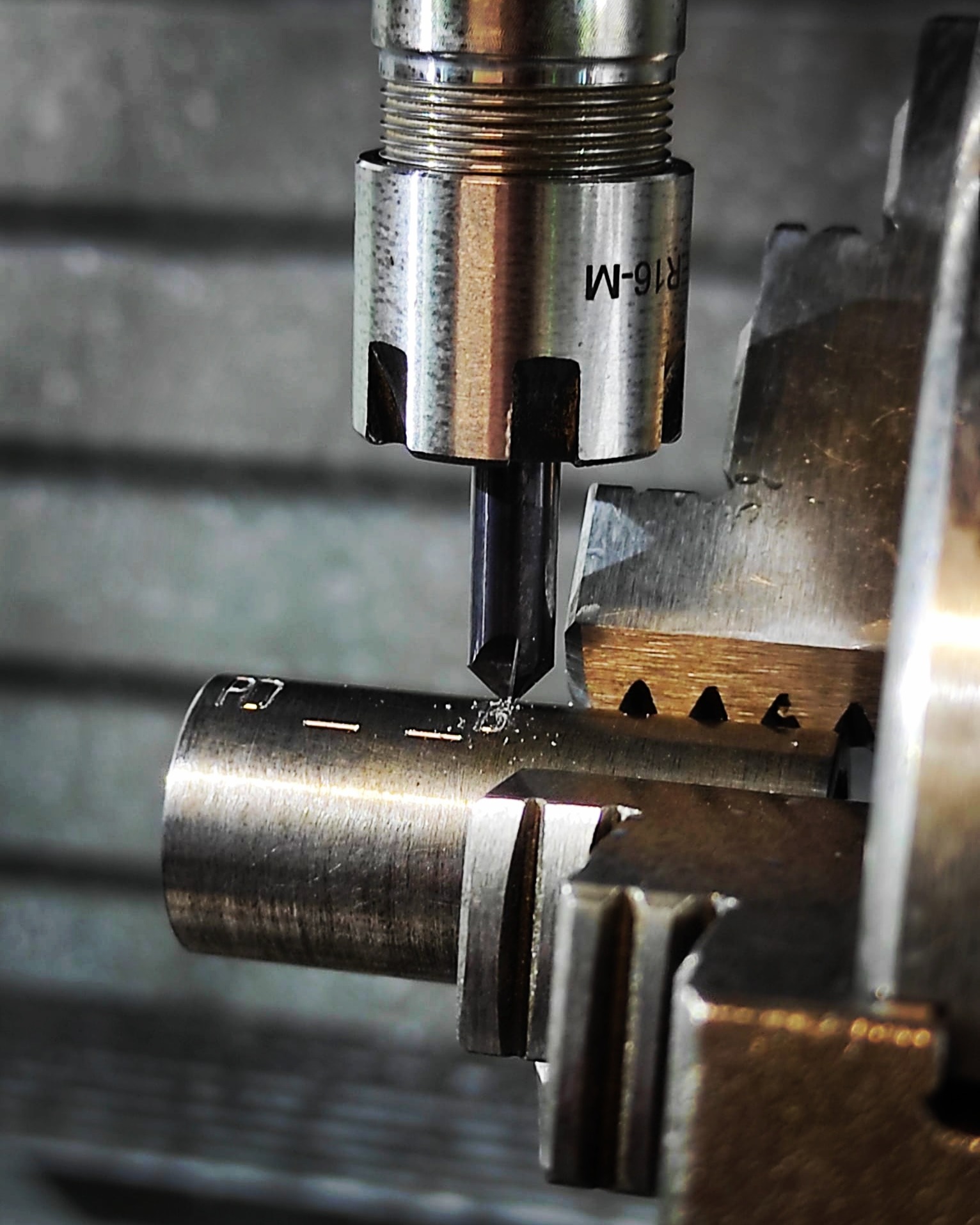 Precyzyjna obróbka metalu: frezarka CNC w trakcie pracy, wióry metalu widoczne wokół ostrza, uchwyt narzędziowy z oznaczeniem 'ER16-M', obrabiany element cylindryczny z naniesionymi oznaczeniami.