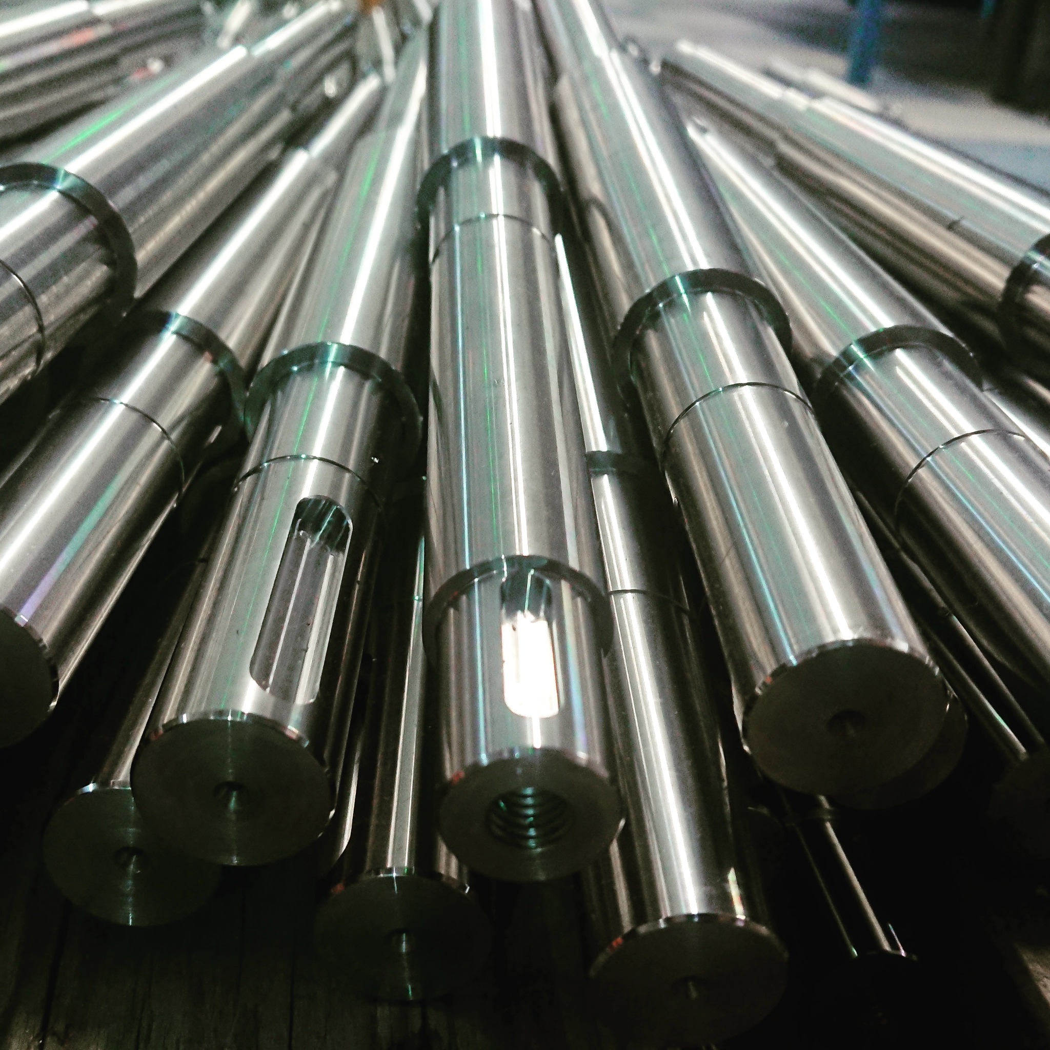Stos polerowanych, metalowych wałków z wyfrezowanymi otworami i gwintowanymi końcówkami, ułożonych pod kątem, w tle widoczna hala produkcyjna.