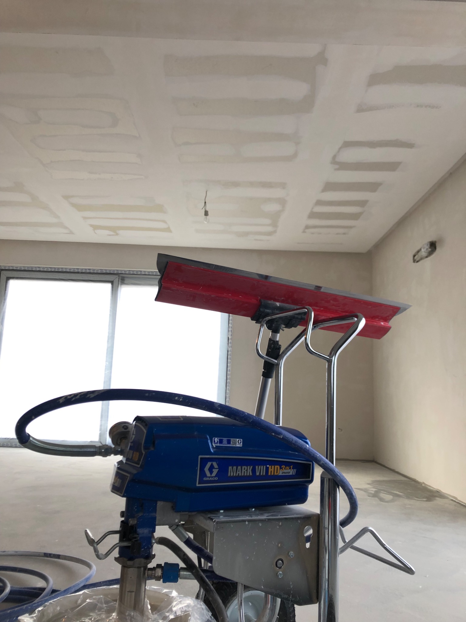 Nierówna powierzchnia sufitu po nałożeniu pierwszej warstwy masy szpachlowej, widoczny agregat malarski Graco Mark VII HD 3in1 z czerwoną szpachlą. Pomieszczenie w trakcie prac wykończeniowych,...