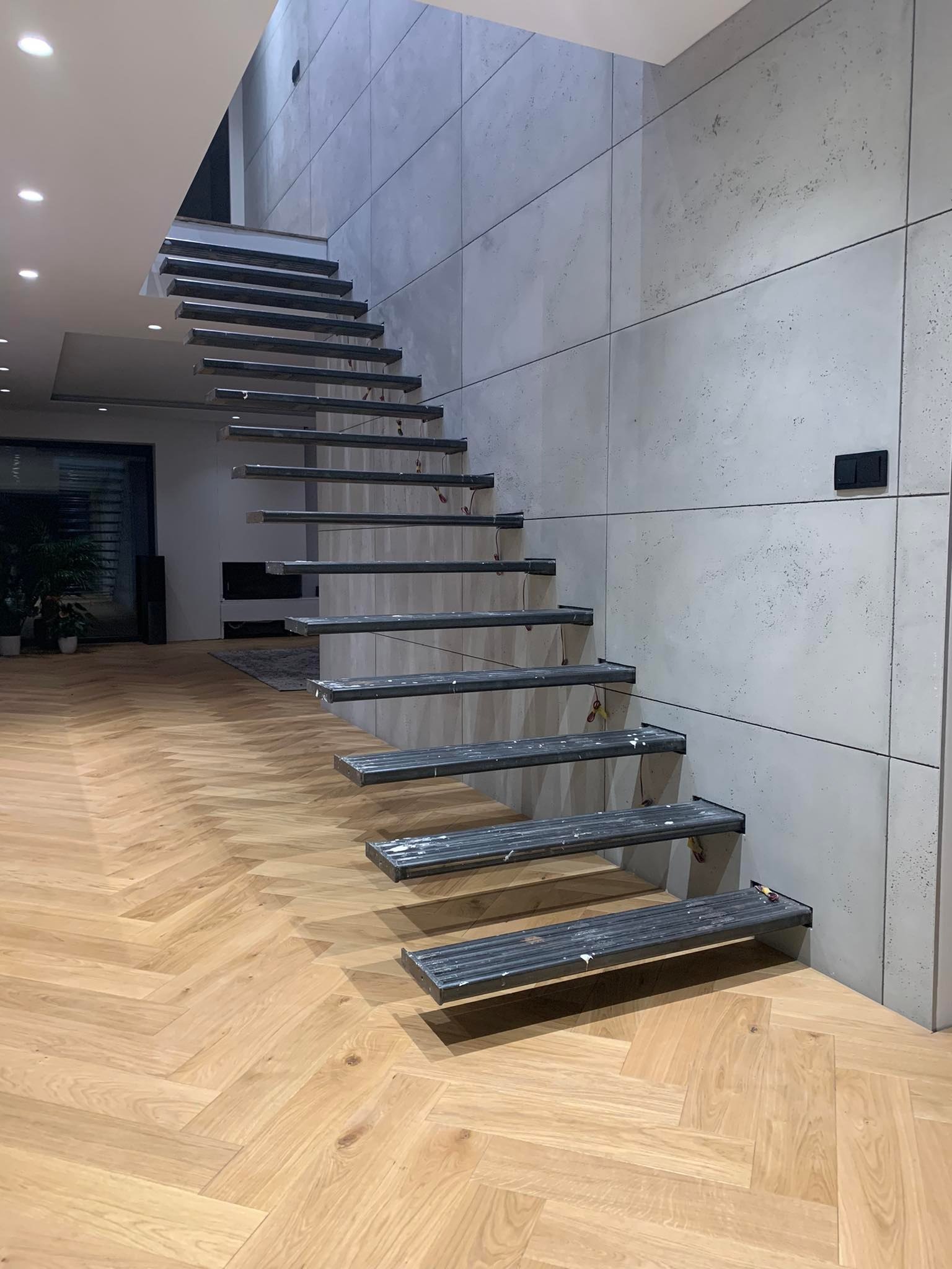 Nowoczesne, ażurowe schody metalowe z przymocowanymi stopniami, zamontowane przy ścianie z betonowych płyt w salonie z parkietem w jodełkę.